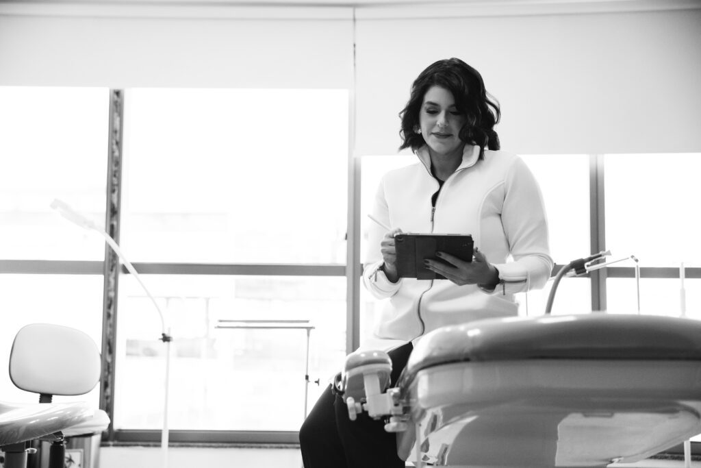 Dra Catarina Almeida com jaleco branco sentada em consultório, segurando um tablet e sorrindo.