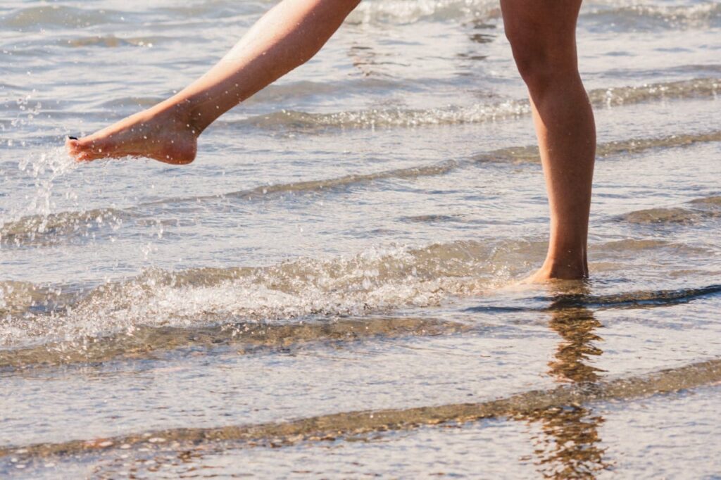 Pernas de uma pessoa caminhando na beira do mar, com água espirrando ao redor.