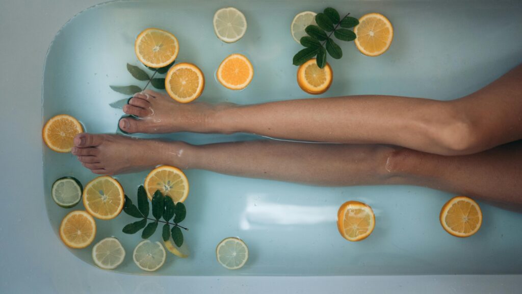 Mulher relaxando em banheira com água e rodelas de laranja e limão, junto a folhas verdes, apenas com as pernas aparecendo. 