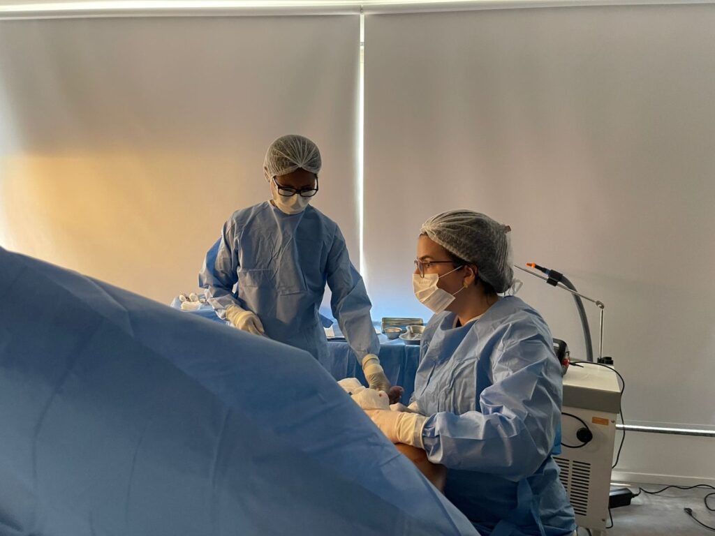 Médicas realizam procedimento cirúrgico em ambiente clínico, utilizando roupas estéreis, toucas e máscaras. 