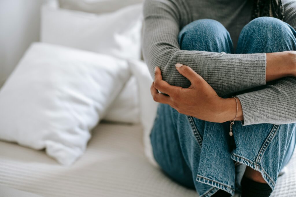 Pessoa sentada em uma cama, com os joelhos dobrados e abraçados junto ao peito, vestindo blusa de manga comprida cinza e calça jeans. 