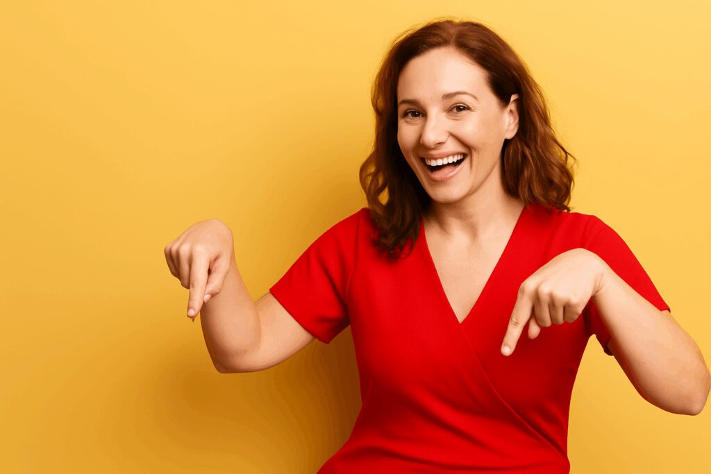 Mulher sorridente vestida de vermelho apontando para baixo com as duas mãos, em frente a um fundo amarelo.