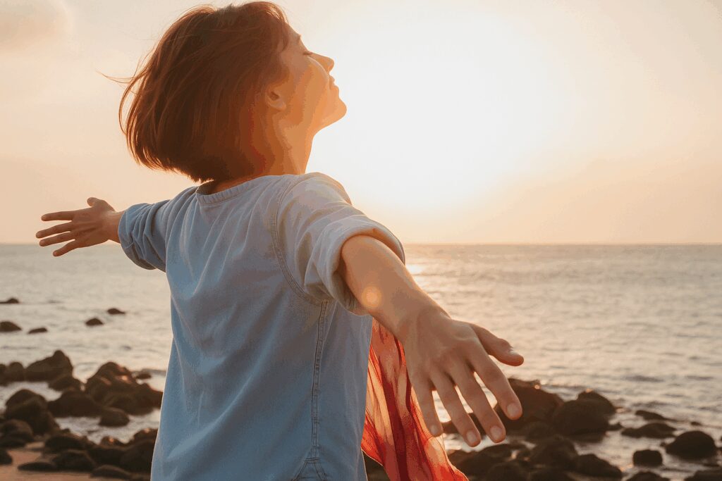 Mulher jovem de braços abertos em uma praia ao pôr do sol, com expressão serena e cabelos ao vento. Ela veste uma camisa jeans clara e segura um lenço vermelho, enquanto observa o mar em meio às pedras, banhada pela luz dourada do fim de tarde.