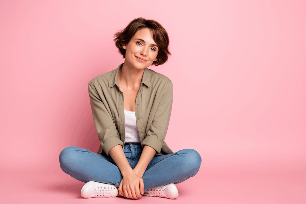 Mulher jovem sentada no chão com as pernas cruzadas, em frente a um fundo rosa, com expressão positiva