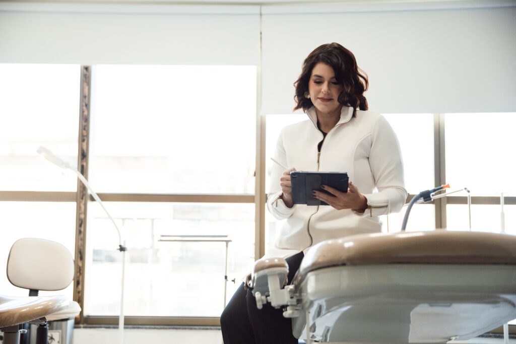 Dra Catarina vestindo jaleco branco, sentada em uma maca de atendimento médico enquanto manuseia um tablet, no seu consultório.