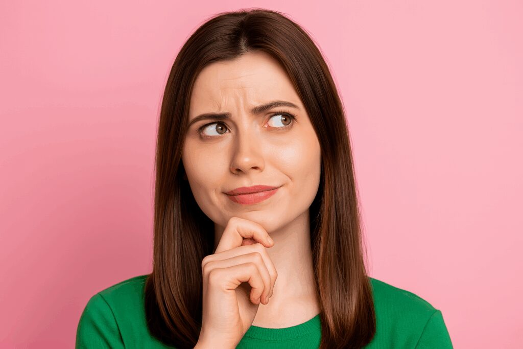 Mulher de cabelos castanhos e lisos, usando blusa verde, posa com expressão pensativa e olhar voltado para cima, diante de um fundo rosa uniforme.