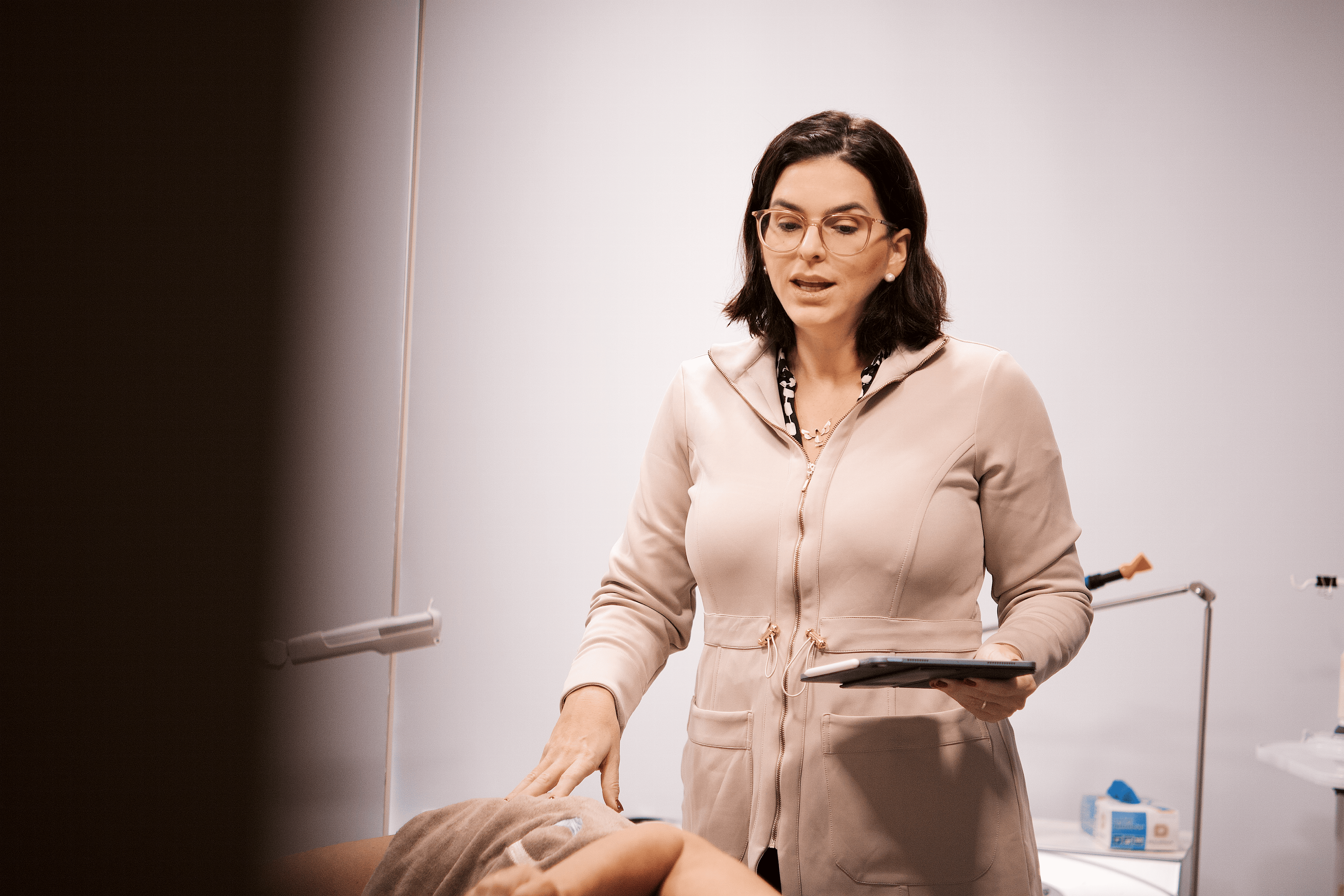 Médica vascular em consultório avaliando paciente durante atendimento, com tablet em mãos.