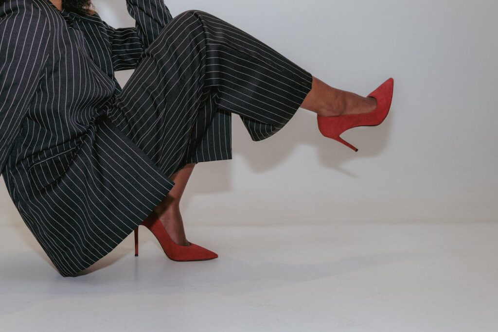 Pessoa usando calça social preta listrada e sapatos de salto alto vermelhos, em pose sentada inclinada.