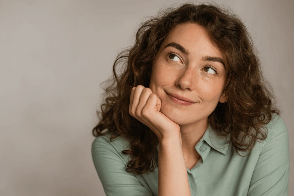 Mulher jovem de cabelos cacheados castanhos apoia o rosto na mão e olha para cima com expressão pensativa e leve sorriso, em fundo neutro.
