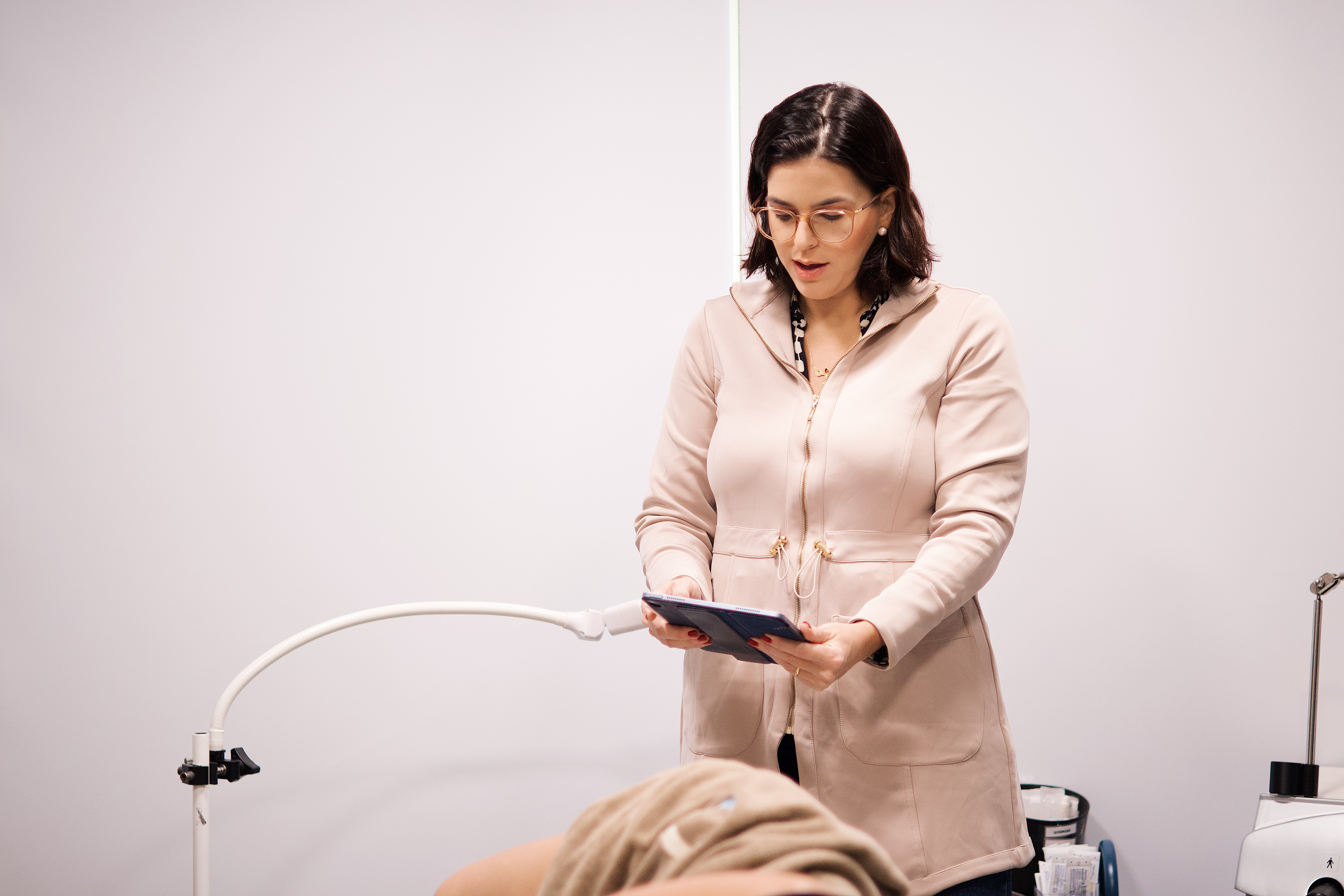Dra Catarina em consultório segurando um tablet durante atendimento, ao lado de equipamento clínico e paciente deitado.