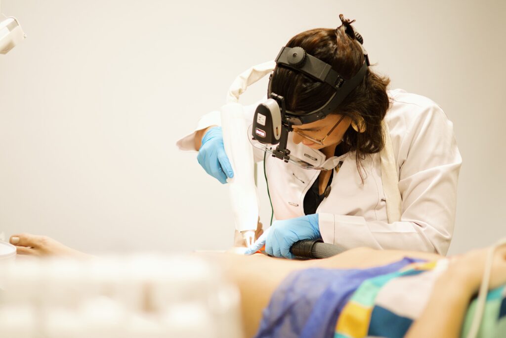 Dra Catarina realizando procedimento a laser na perna de um paciente, utilizando equipamentos de proteção e tecnologia especializada.
