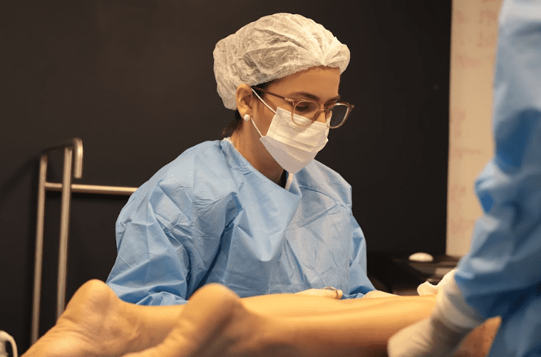 Médica vascular usando touca, máscara e jaleco estéril realiza procedimento nas pernas de um paciente em ambiente clínico.
