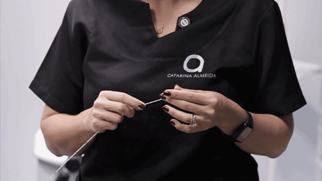 Dra Catarina vestindo jaleco preto com o nome "Catarina Almeida" bordado, segura delicadamente uma fibra fina de endolaser com ambas as mãos.