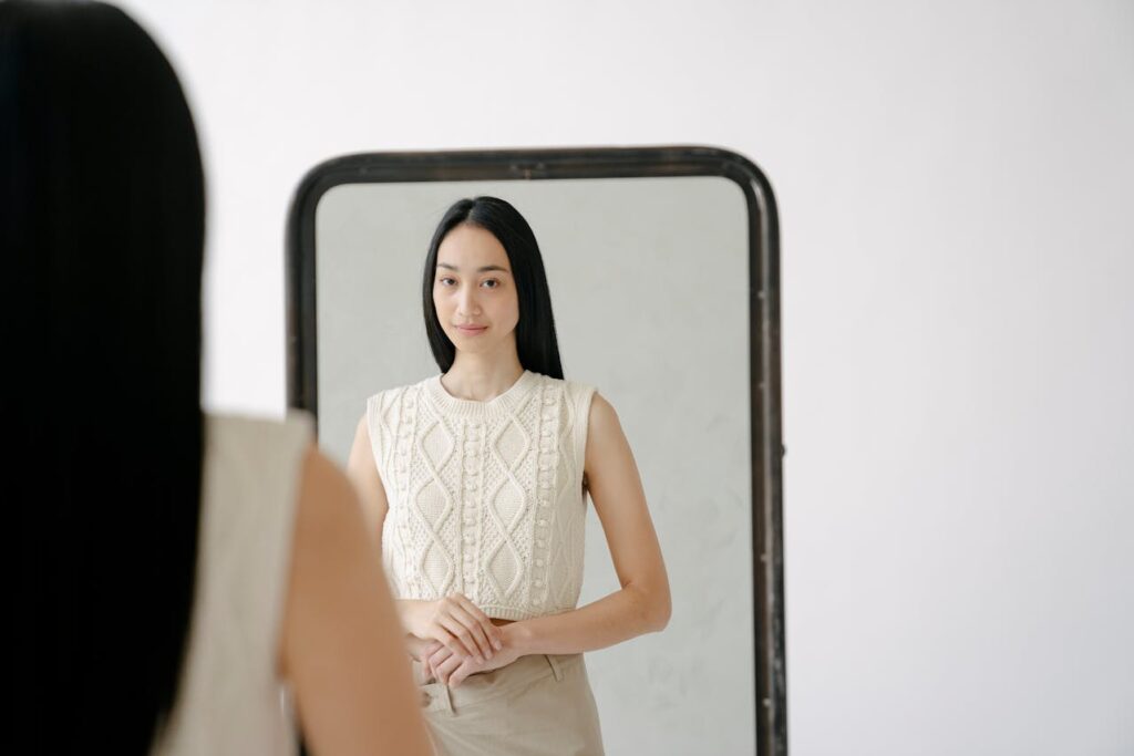 Mulher de cabelos longos e lisos observa seu reflexo em um espelho grande, com expressão serena e leve sorriso, vestindo blusa clara de tricô e calça bege